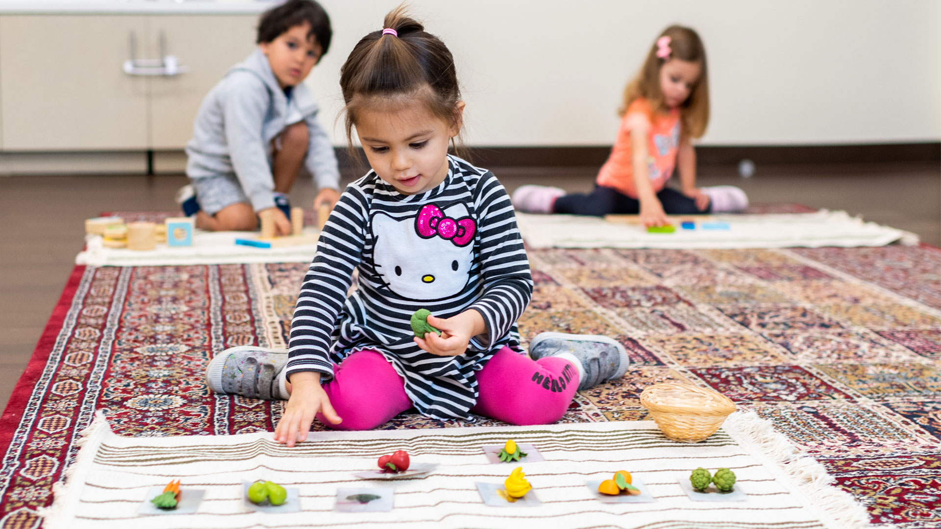 A little girl wearing a striped dress placing toy fruits on her white rug